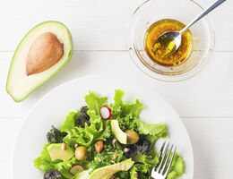 A bowl of oil-based dressing next to a plate of salad and half an avocado.