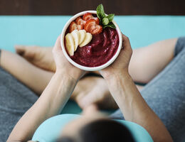 A woman, seen from above, sits cross-legged while holding a smoothie bowl topped with strawberry and banana slices.