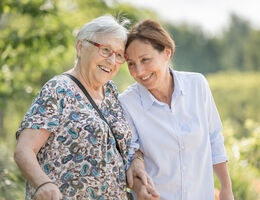 An older woman and a younger woman stand arm in arm and lean their heads together. 