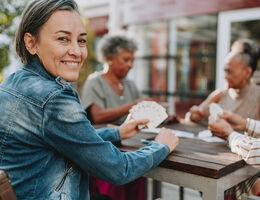 A woman smiles at the camera as she plays cards outdoors with other older women.