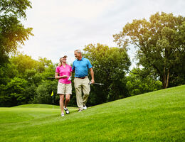A man and woman smile as each carries a golf club on a golf course.