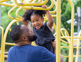 A dad helps his young daughter do the monkey bars on a playground.